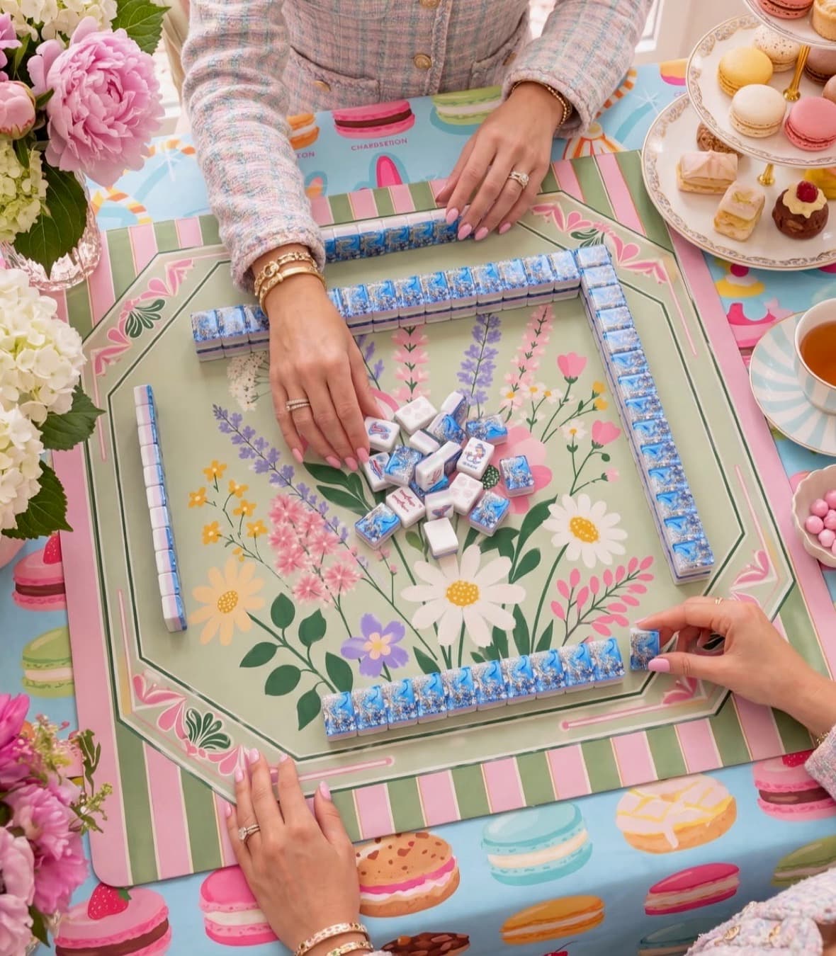 Two players arranging mahjong tiles at a floral table set with tea and pastries